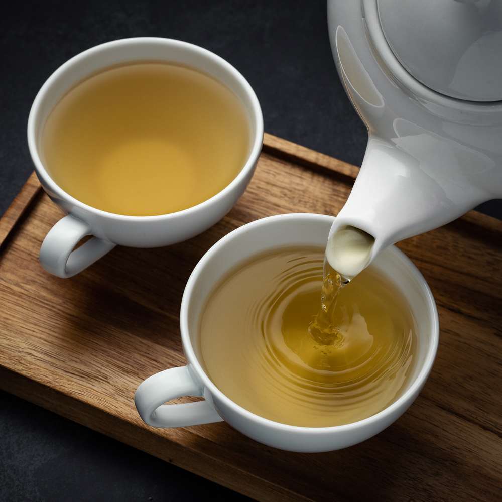 Two cups of tea on a wooden tray, one has yellow tea being poured into it from a tea pot