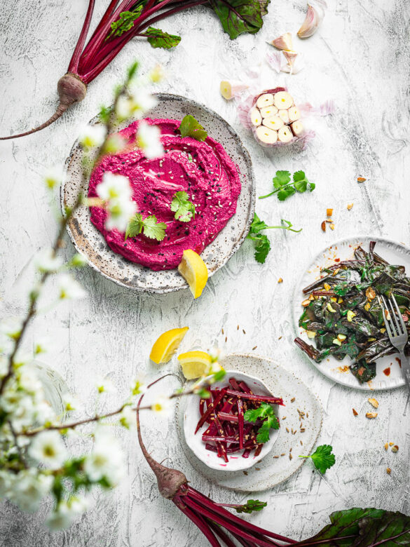 Overhead scene on white with a plate of beetroot hummus, pickled beetroot stems, and a plate with sautéed beetroot greens