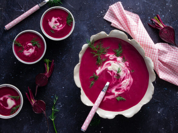 Overhead shot of a large and two small bowls of pink beetroot soup with sour cream and dill garnish, plus pink serviette and spoons