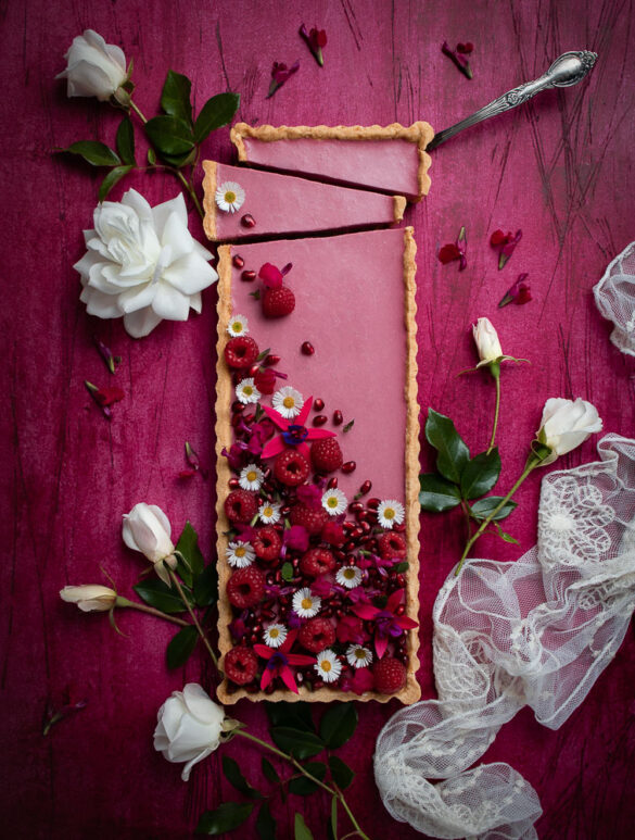 A very pretty pink scene of a pink, rectangular berry tart, with white flowers and raspberries