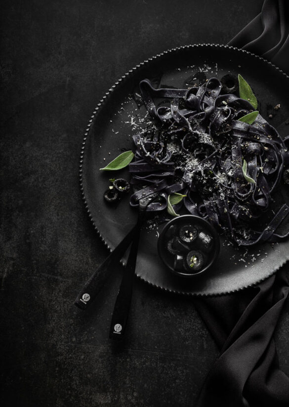 Black pasta on a black plate with black cutlery, black olives, on a black background