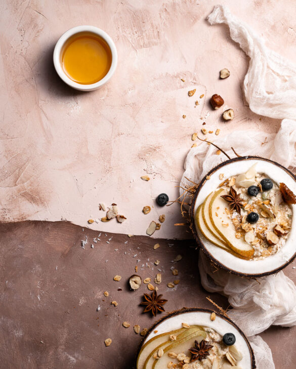 A top down scene like a painting, with Bircher muesli in two coconut halves, a sheer scarf, and a little bowl of honey