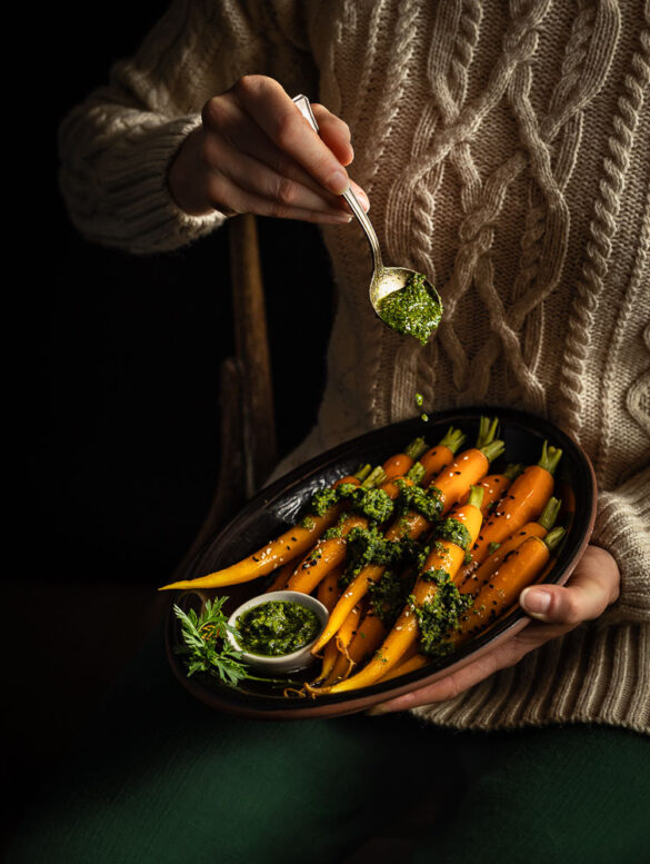 A person sitting down with a plate of steamed carrots, drizzling green carrot top pesto over the carrots