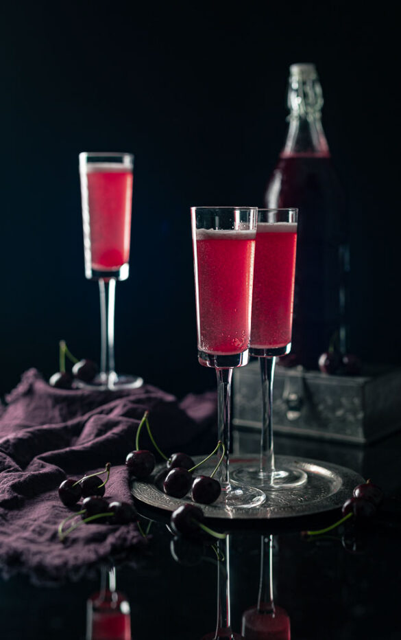 Moody scene with three tall champagne glasses of cherry cordial and a bottle in the background
