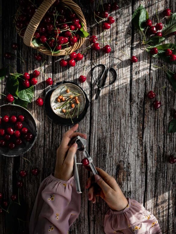 A rustic table scene with a basket of cherries and two hands pitting the cherris