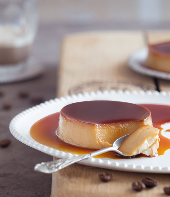 A white plate with creme caramel with a spoon, with another plate and coffee in the background
