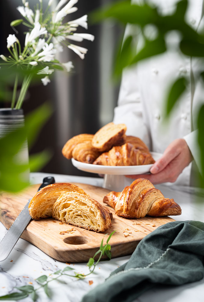 A person in a chef jacket placing a cake stand with freshly baked croissants onto a table, with a cut open croissant in the foreground