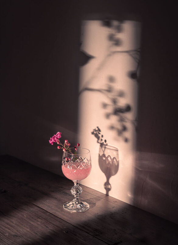 A crystal glass with a pink drink and a flower garnish, lit by light shining through a door, casting a shadow on the wall