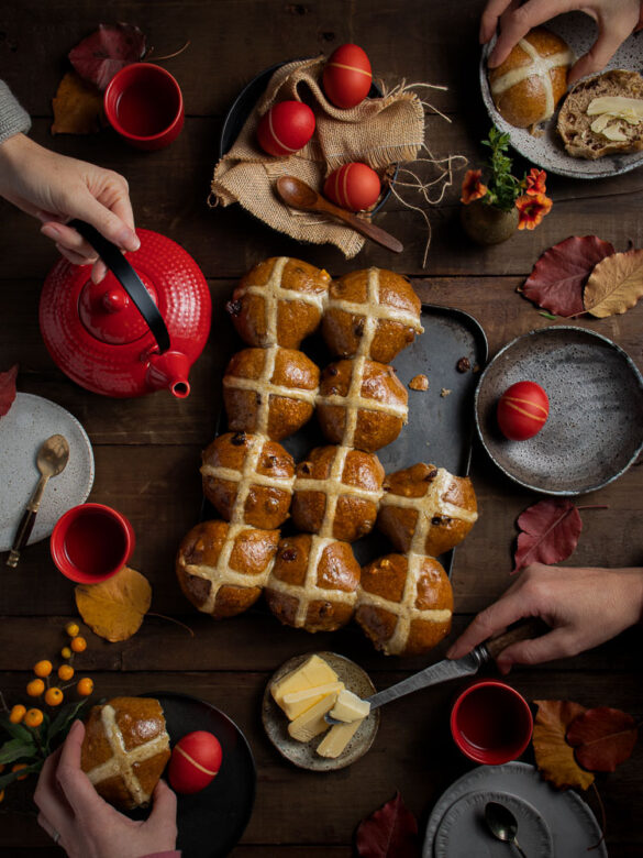 Homemade hot cross buns on a table and various hands helping themselves to buns and tea and butter