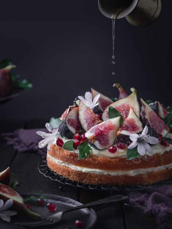 A beautifully decorated two-layer cake with fresh figs, pomegranate seeds, and honey being drizzled onto the cake