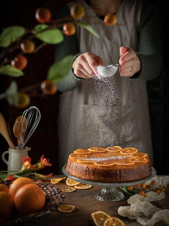 Flourless orange cake decorated with orange slices and a person sprinkling icing sugar on top
