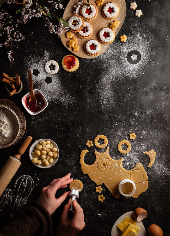 A baking scene showing flower shaped cookies being cut out of dough and filled with jam