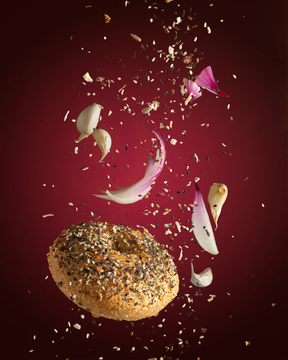 An everything bagel flying through the air with seeds, garlic cloves and onion wedges