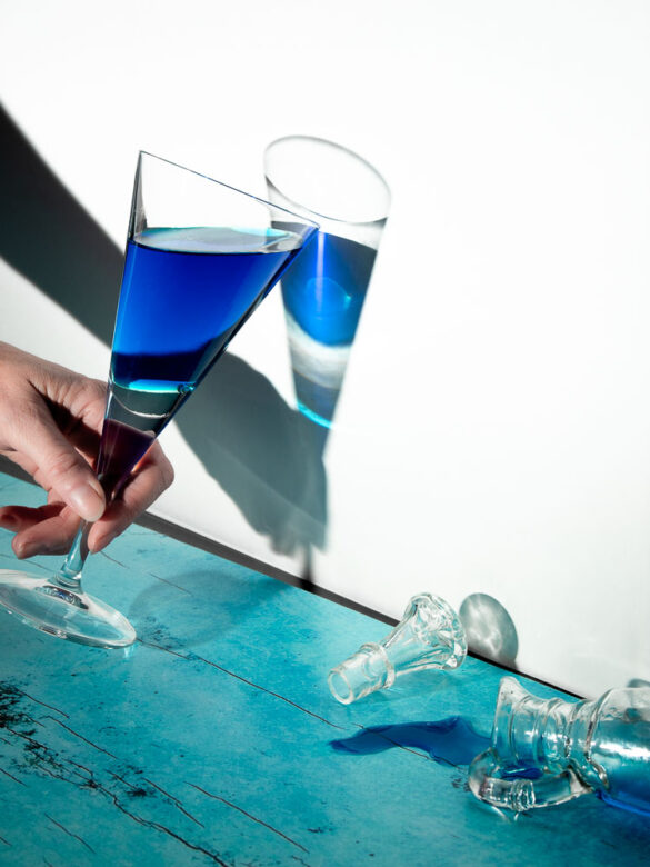 A hand holding a glass with a blue drink casting a mirroring shadow on a white wall