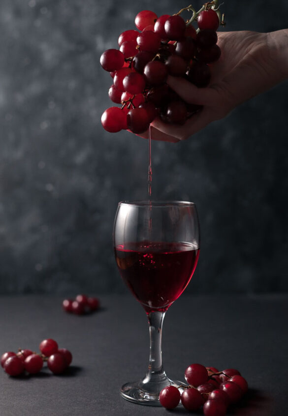 A hand squeezing juice from a bunch of red grapes into a wine glass