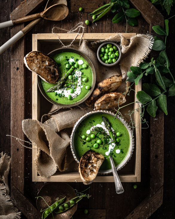 two bowls of green pea and asparagus soup in a wooden book with crusty bread and cream