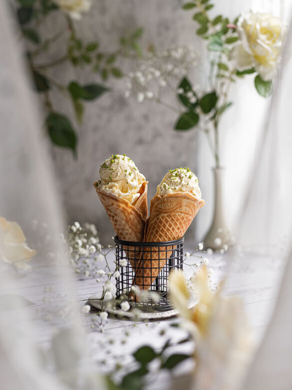 A peak through sheer curtains onto a table scene by a window with two cones of ice cream standing up in a V formation