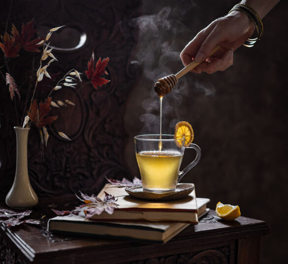 Steaming hot lemon tea with a hand holding a honey dipper dripping some honey into the liquid
