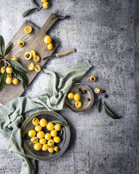 A rustic grey surface with a chopping board, a plate with loquats, a green napkin and a knife with some cut loquats