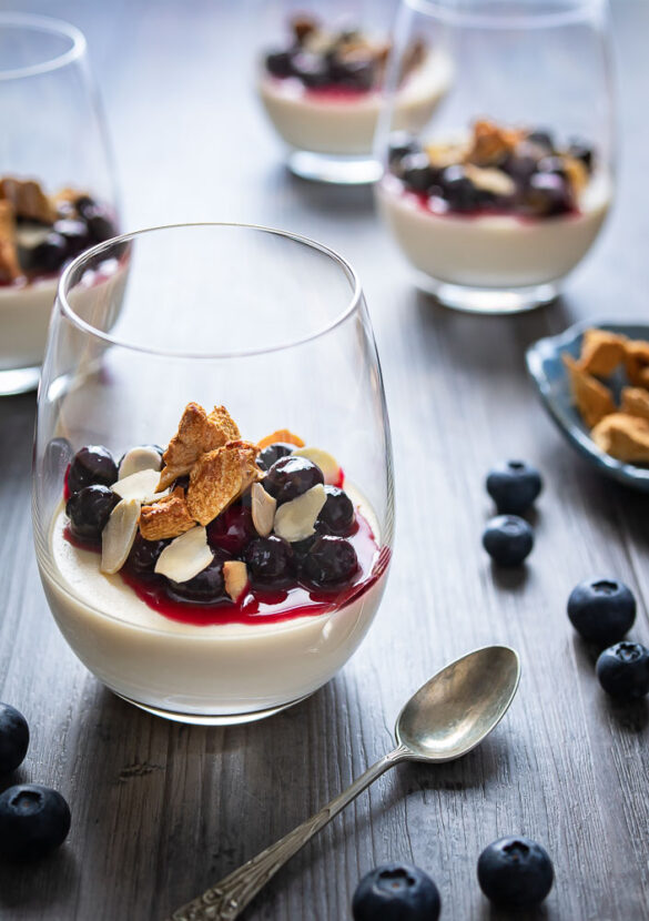 A glass of panna cotta with blueberries and honeycomb crumbled on top, with more glasses blurry in the background