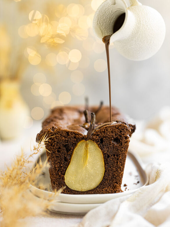 A light and airy scene of a chocolate cake, cut open to reveal an in-baked pear, being drizzled with chocolate sauce