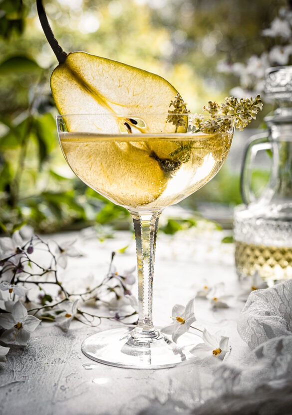 A garden table scene with a close up of a coupe glass with cider and a slice of pear