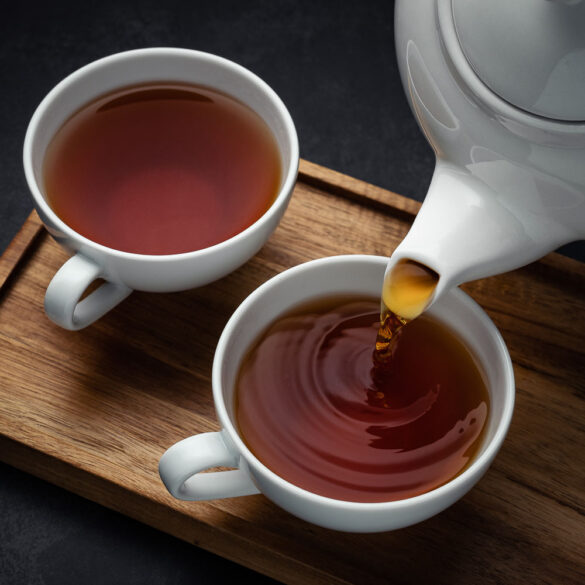 Two cups of tea on a wooden tray, one has black tea being poured into it from a tea pot