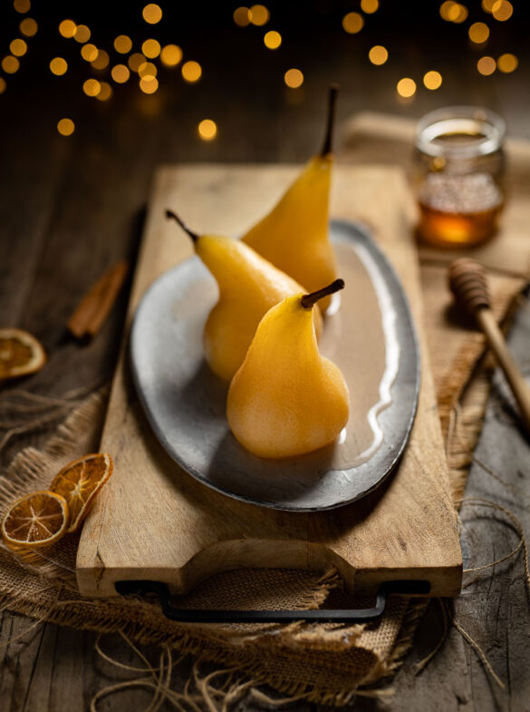 A festive looking scene of three golden poached pears lined up on a plate, with golden fair lights, honey and dried oranges