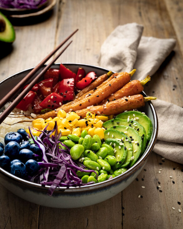 A poke bowl with red capsicum, orange carrots, yellow corn, green avocado and edamame, purple cabbage, and blueberries, like a rainbow