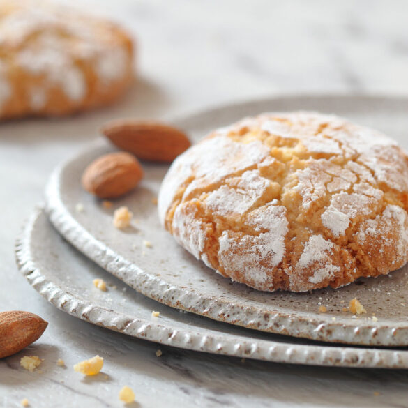Close up of a ricciarelli biscuit on two white rustic plates, with a few almonds sprinkled around