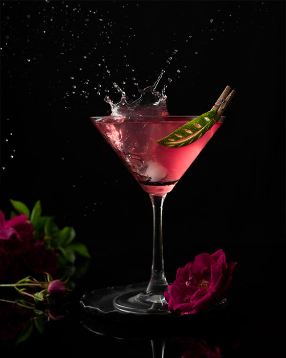 A glass of Cosmo, glowing pink, with a big splash, lime garnish and large pink flowers in front of a black background