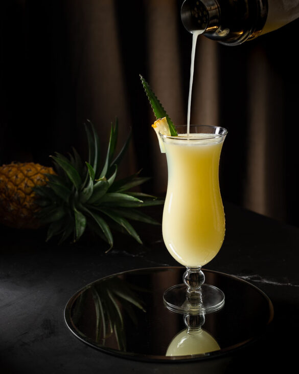 A glass of yellow pina colada on a mirror, with liquid being poured into the glass, a pineapple in the background and pineapple garnish