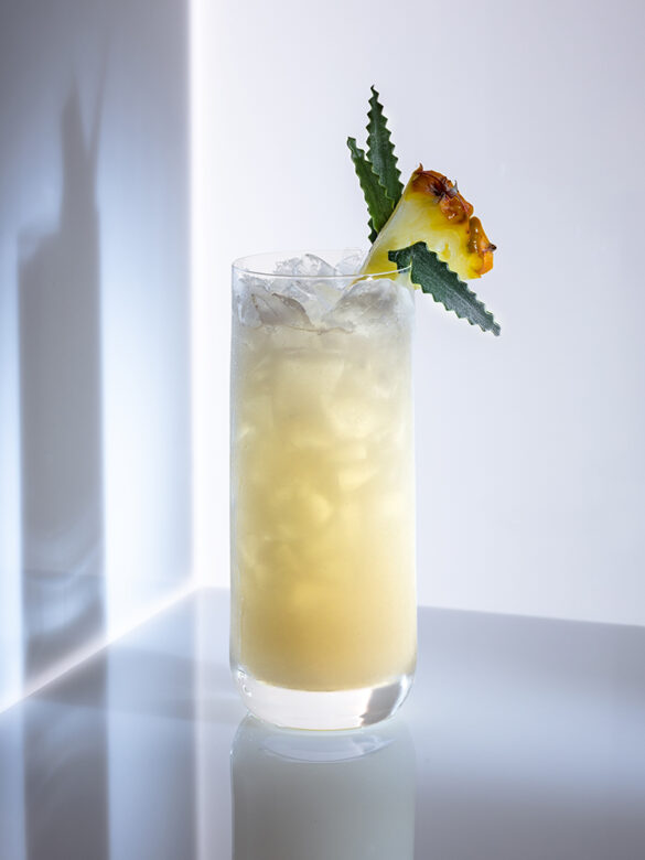 A pina colada in a tall glass with pineapple garnish, casting a shadow onto the white wall