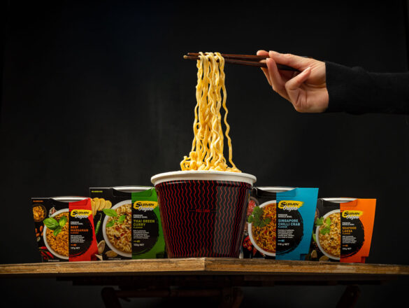 Four cups of Suimin noodles in a V position on a table corner, with a cup of noodles in the middle and a hand lifting out some noodles with chopsticks