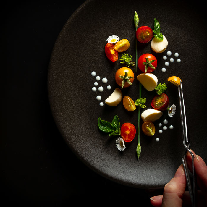 Overhead shot of a deconstructed caprese salad with a hand placing a tiny tomato with pliers