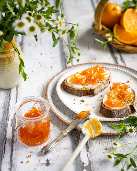 Two slices of rye sourdough with orange marmalade, daisies in a vase, and oranges in a basket